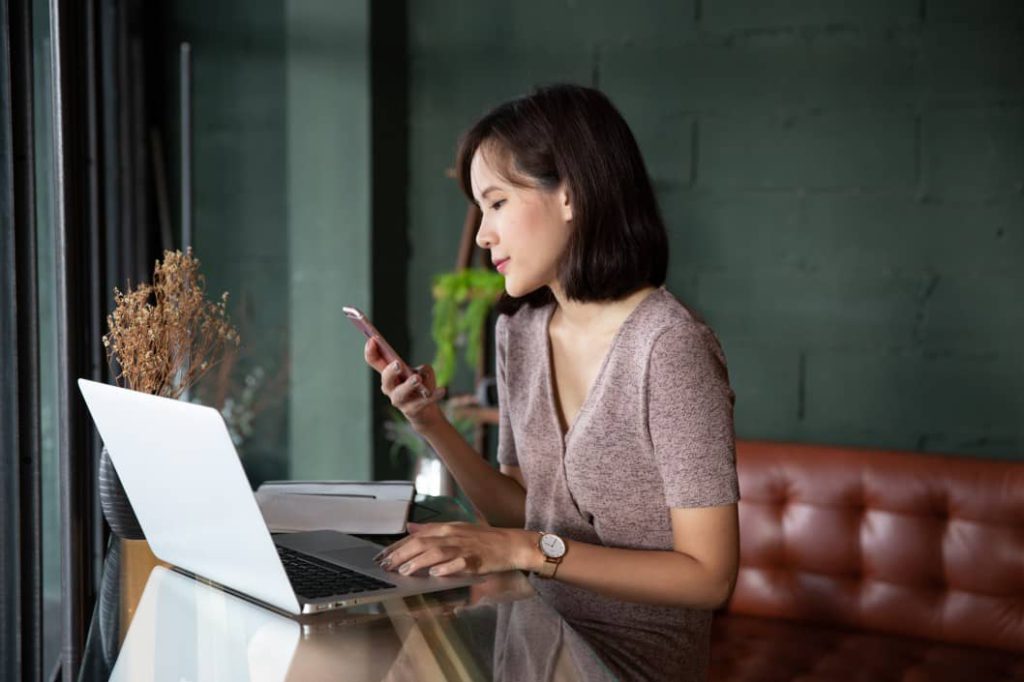 Young beautiful asian woman working in loft cafe or office. Holding smart phone and using laptop computer