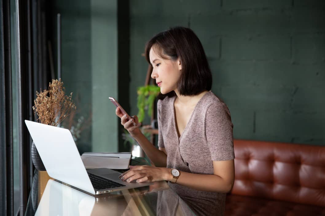 lower cost coworking environment Young beautiful asian woman working in loft cafe or office. Holding smart phone and using laptop computer. business, window, office, table, laptop, work, mobile, notebook, freelance, secretary, technology, tech