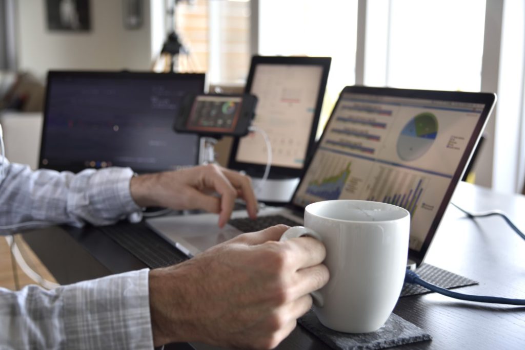 working-with-many-computer-screens-with-data-charts-and-graphs-hand-on-mug-of-coffee-multitasking-at_t20_Rw241X